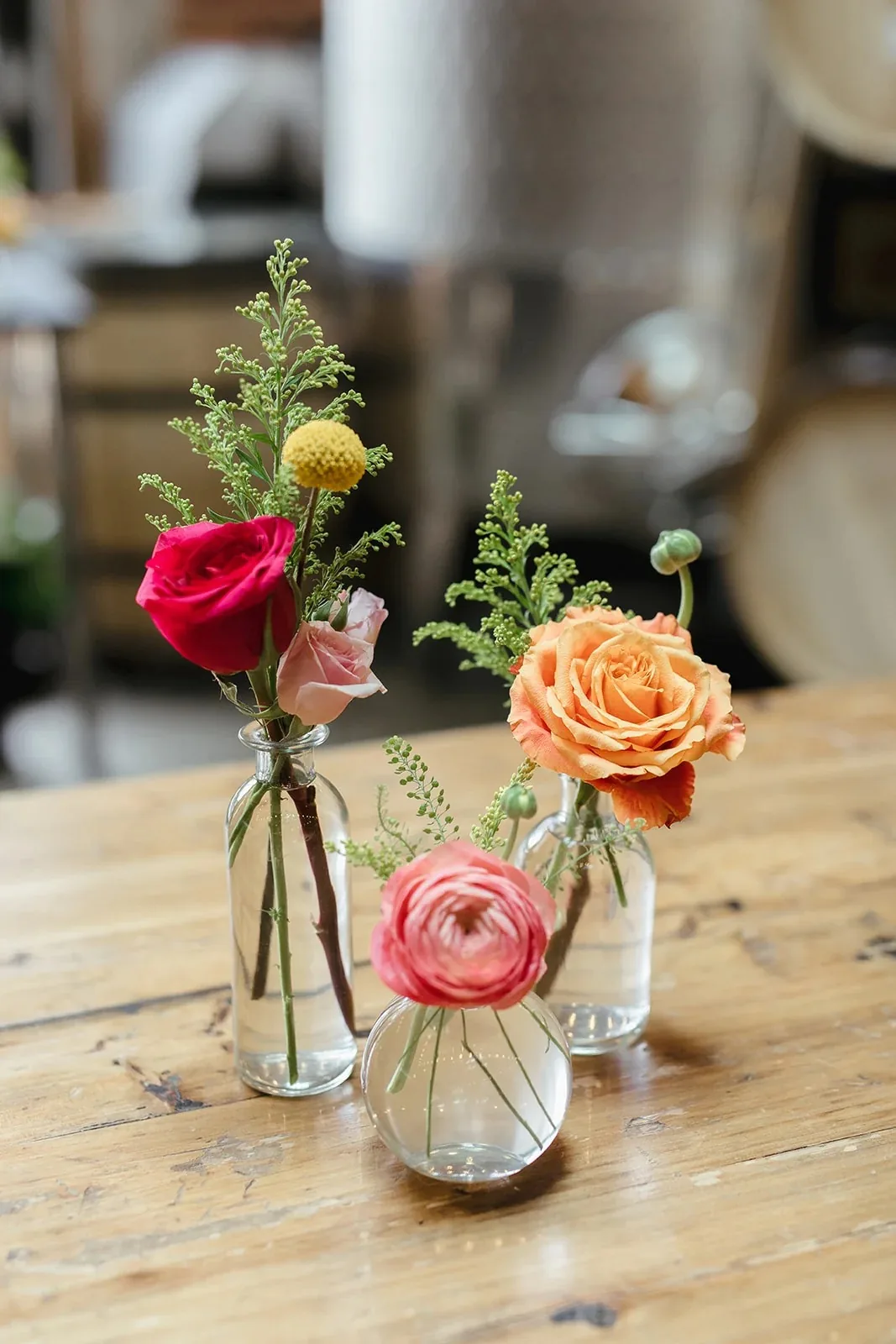 Three clear bud vases with a pink rose, peach rose and coral ranunculus on a wooden reception table